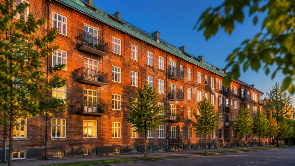 Rød murstensejendom i flere etager med hvide vinduer i aftenlys, hvor enkelte vinduer er tændt