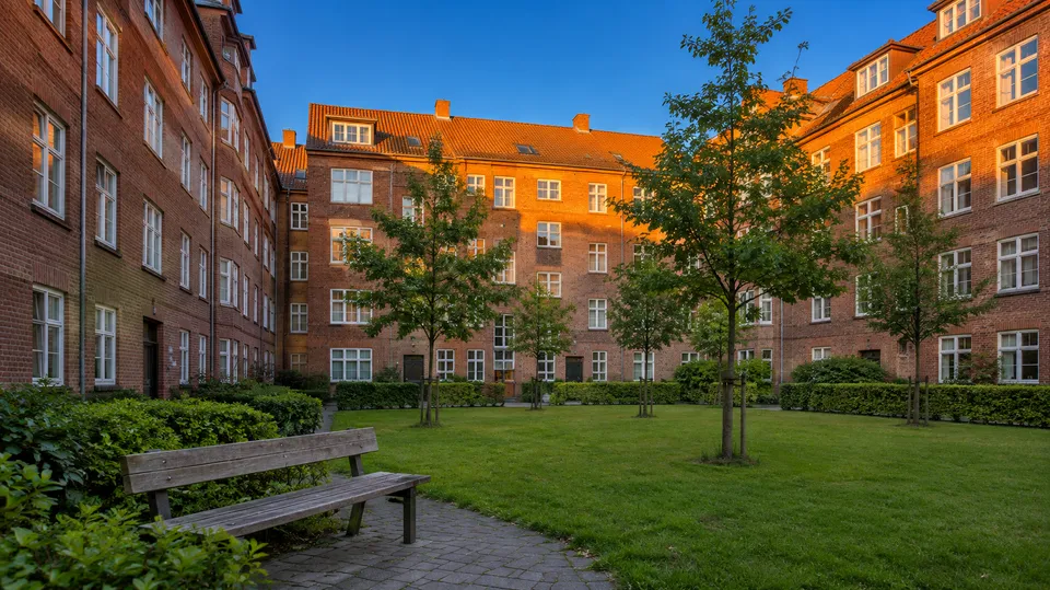 Grøn gårdhave med en bænk omgivet af røde murstensfacader i aftenlys